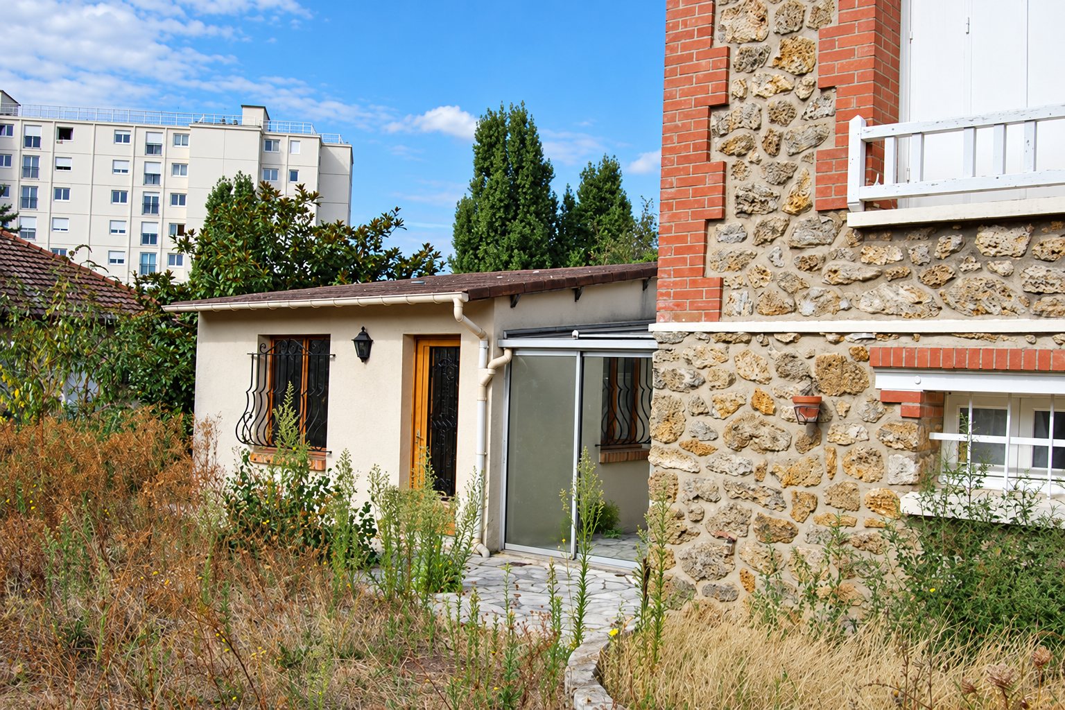 Petite annexe en crépi à côté d’une maison en pierre meulière, jardin en friche